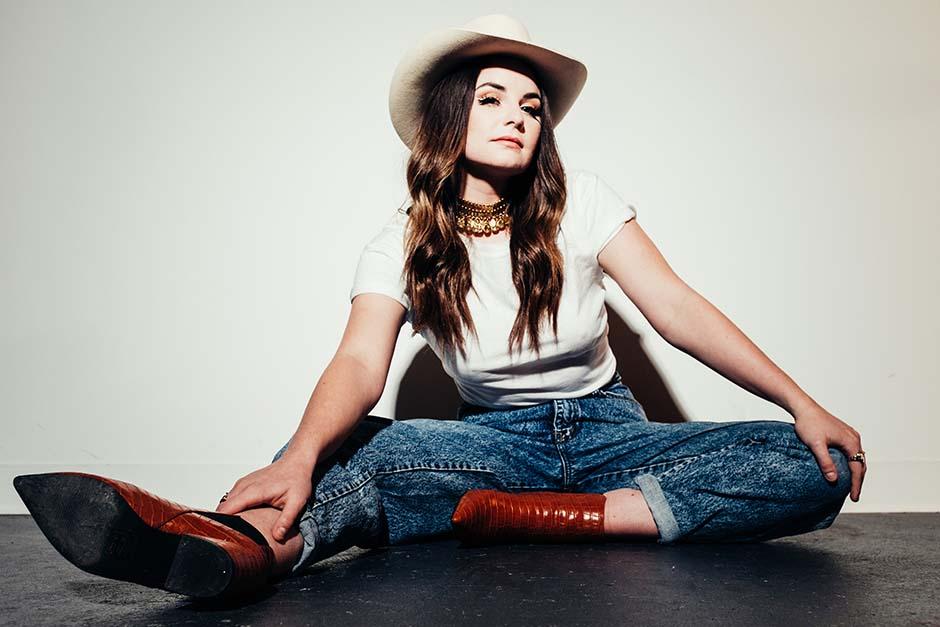 Mariya Stokes is wearing brown boots, blue jeans, white t-shirt, and a white cowboy hat. She is seated on the ground in front of a white wall. 