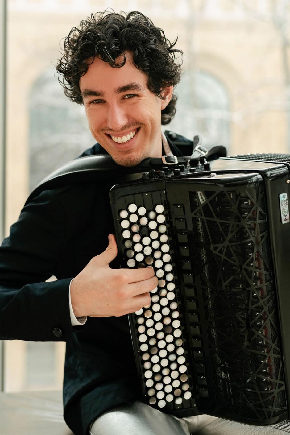 Michael Bridge is playing a black accordion and smiling at the camera