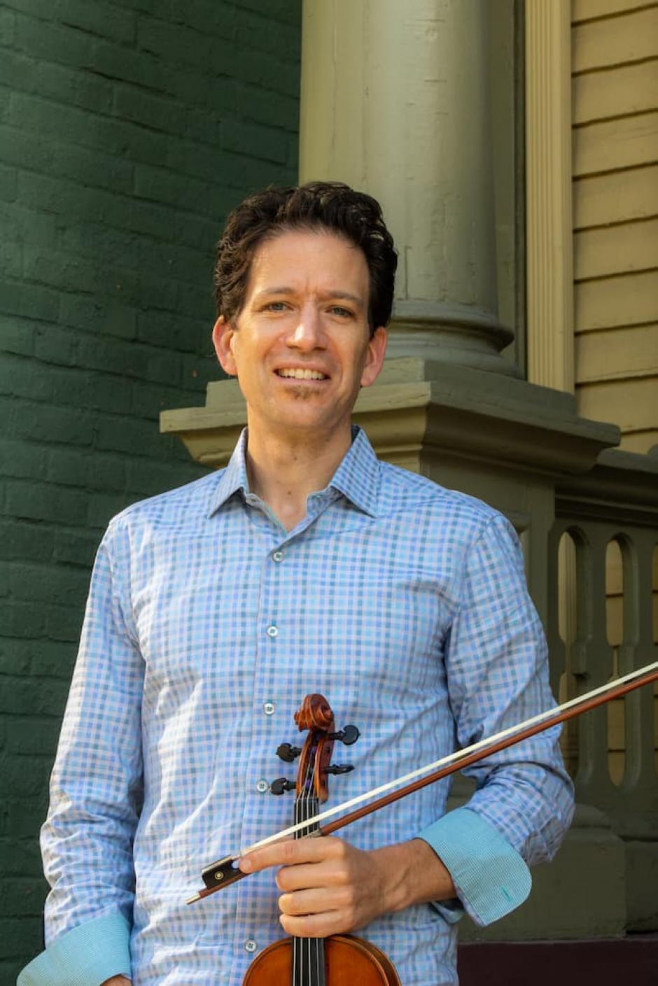 Sebastian Ruth holds a violin and bow while smiling into the camera
