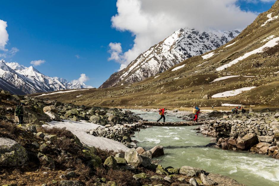 Hikers cross a foot bridge over a small glacial river in the mountains.