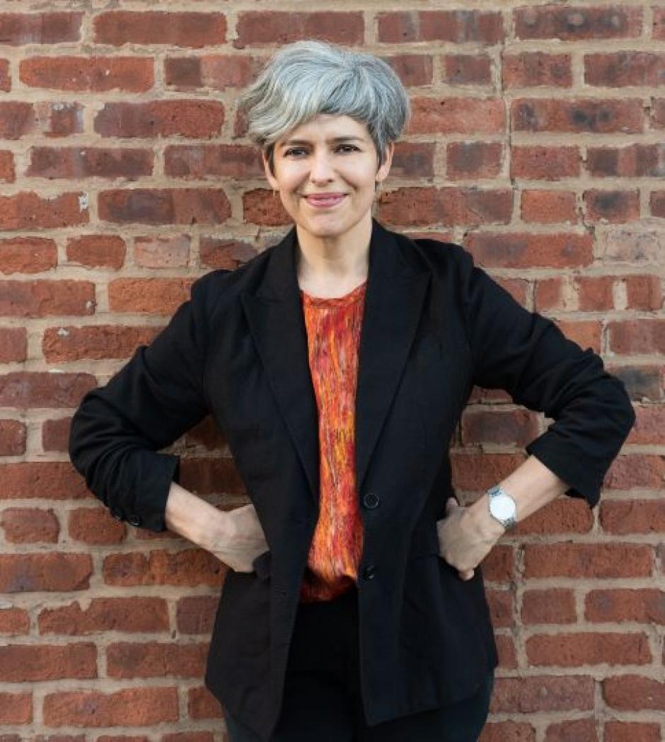Photo of program faculty, Monica de la Torre, standing with hands on hips against a brick wall backdrop