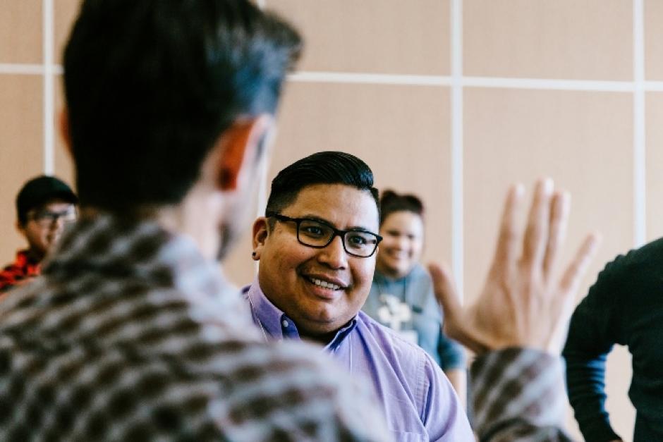 Photo shows Students in Introduction Negotiation Skills Training Course, one student stands to left with back to camera and right hand up and open as an another student looks forward to high five.