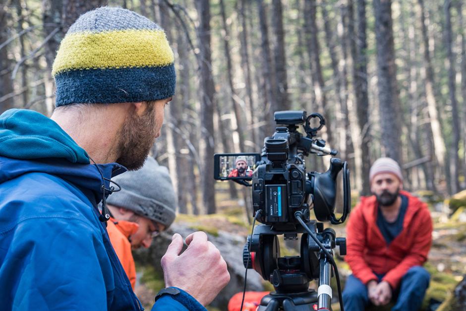 Person wearing a blue jacket and striped hat points video camera at man in red jacket, seated