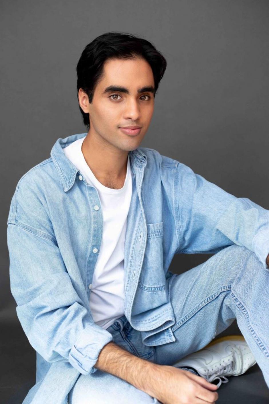Davinder Malhi is wearing a light blue denim shirt unbuttoned over a white tshirt and is smiling into the camera