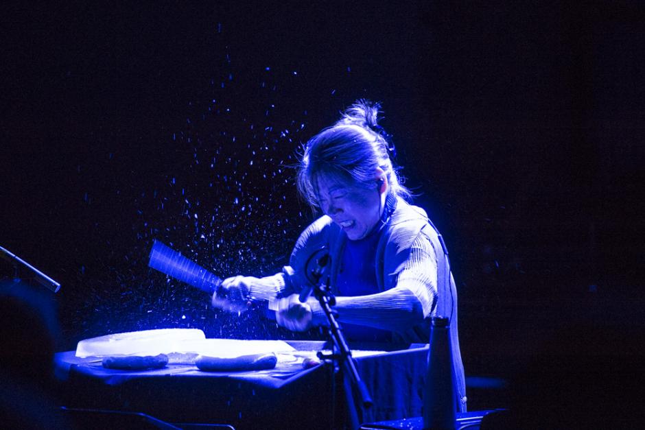 A woman lit with an overhead blue light beats a unique drum set.