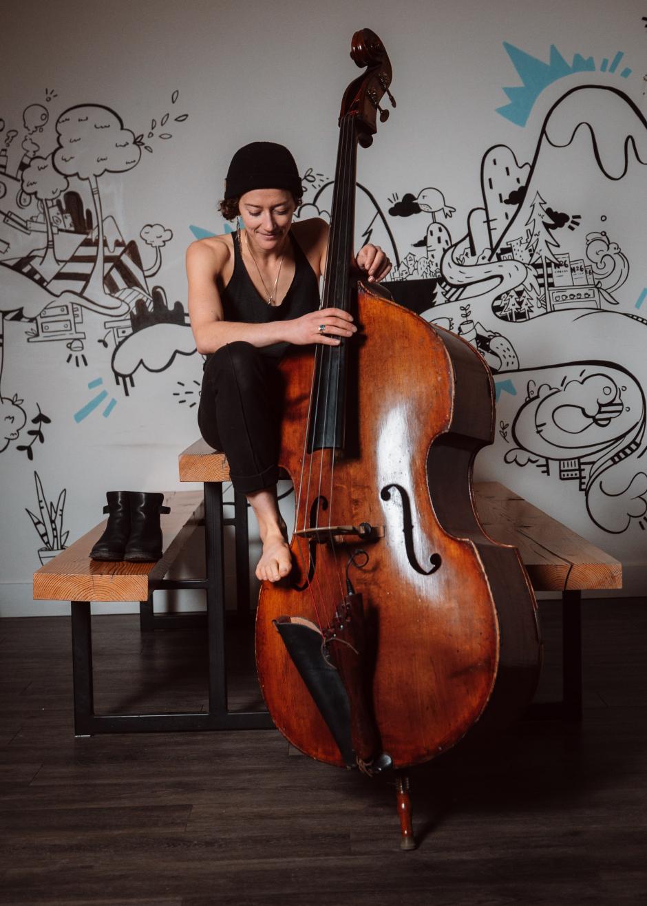 Jill sits in front of a white wall with black drawing art on the wall. She plays an upright base and wears a black toque. 