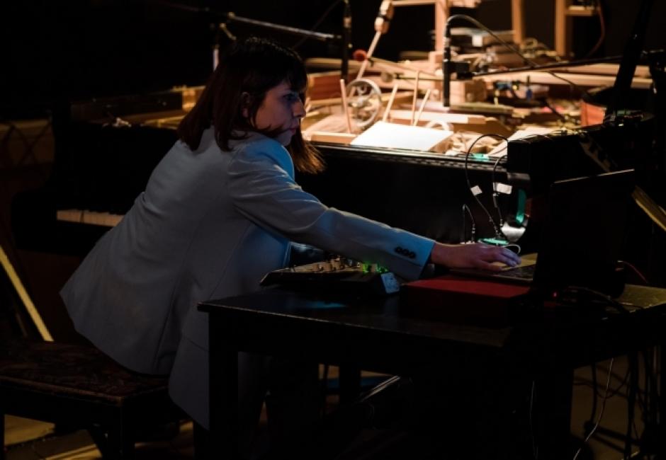 Anoush sits at a piano with additional musical electronic equipment. Anoush wears a grey blazer. There is light on the piano, but Anoush is slightly in shadow.