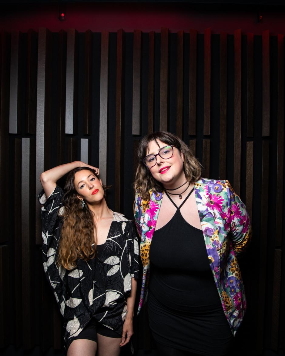 Marie-Clo on the left and éemi on the right pose in front of a striped background. Marie-Clo wears a dark outfit with white decals and éemi wears a black under layer with a colourful blazer. They pose for the camera.