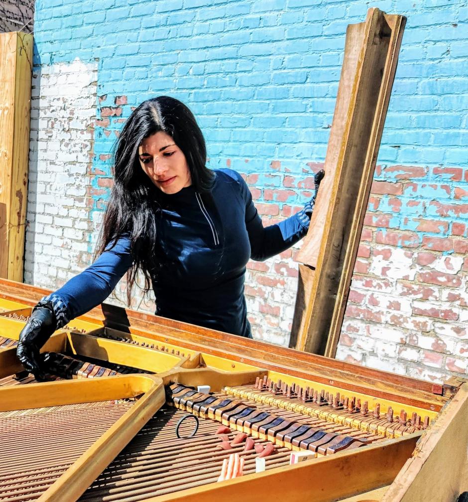 Nicole stands in front of a blue and white rustic brick wall playing the inside of a piano. 