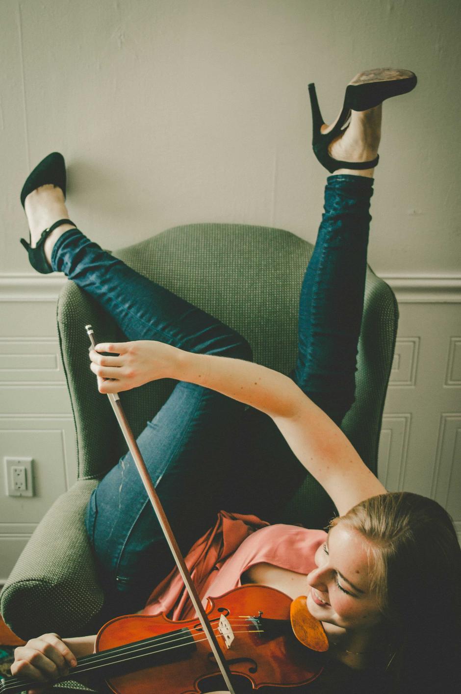 Lauren lays on a chair and plays a violin. She is wearing jeans and smiles.