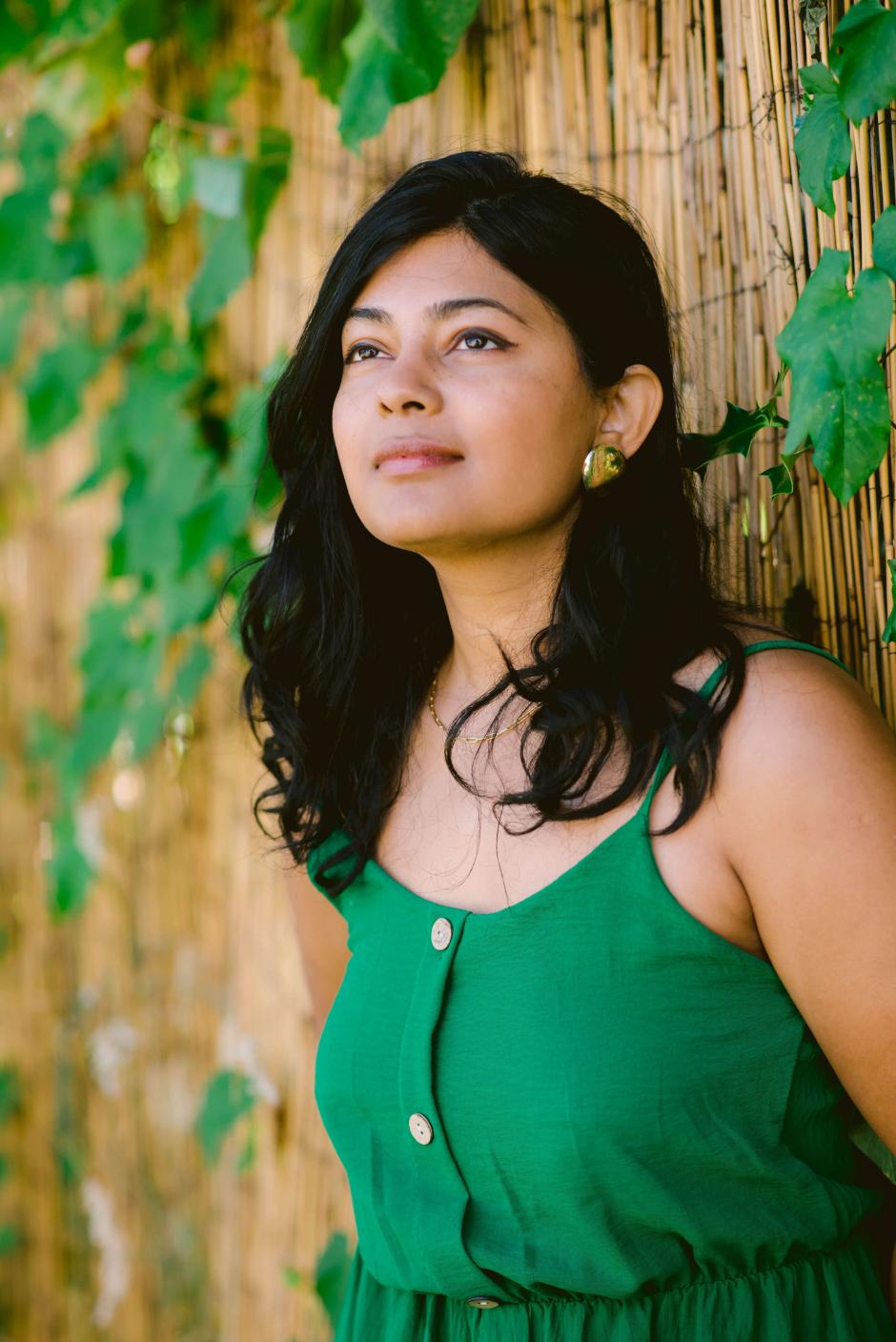 Priyanka is in a green dress against a naturesque and fenced background. She stares off into the left of the frame. 