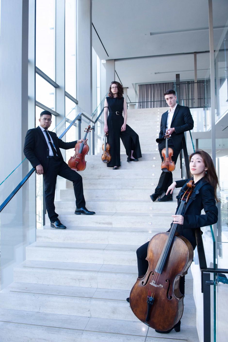 The Dior Quartet stand on white stairs holding their instruments. They lean on the railings and look at the camera. 