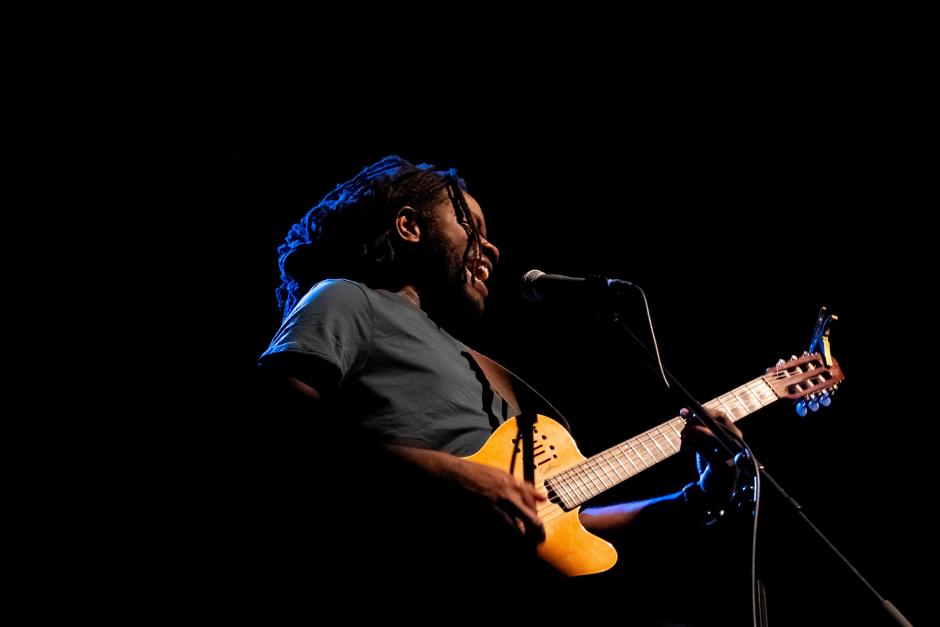 Duane holds a guitar. He is performing and the shot is taken from an angle underneath him. He is singing into a microphone. 