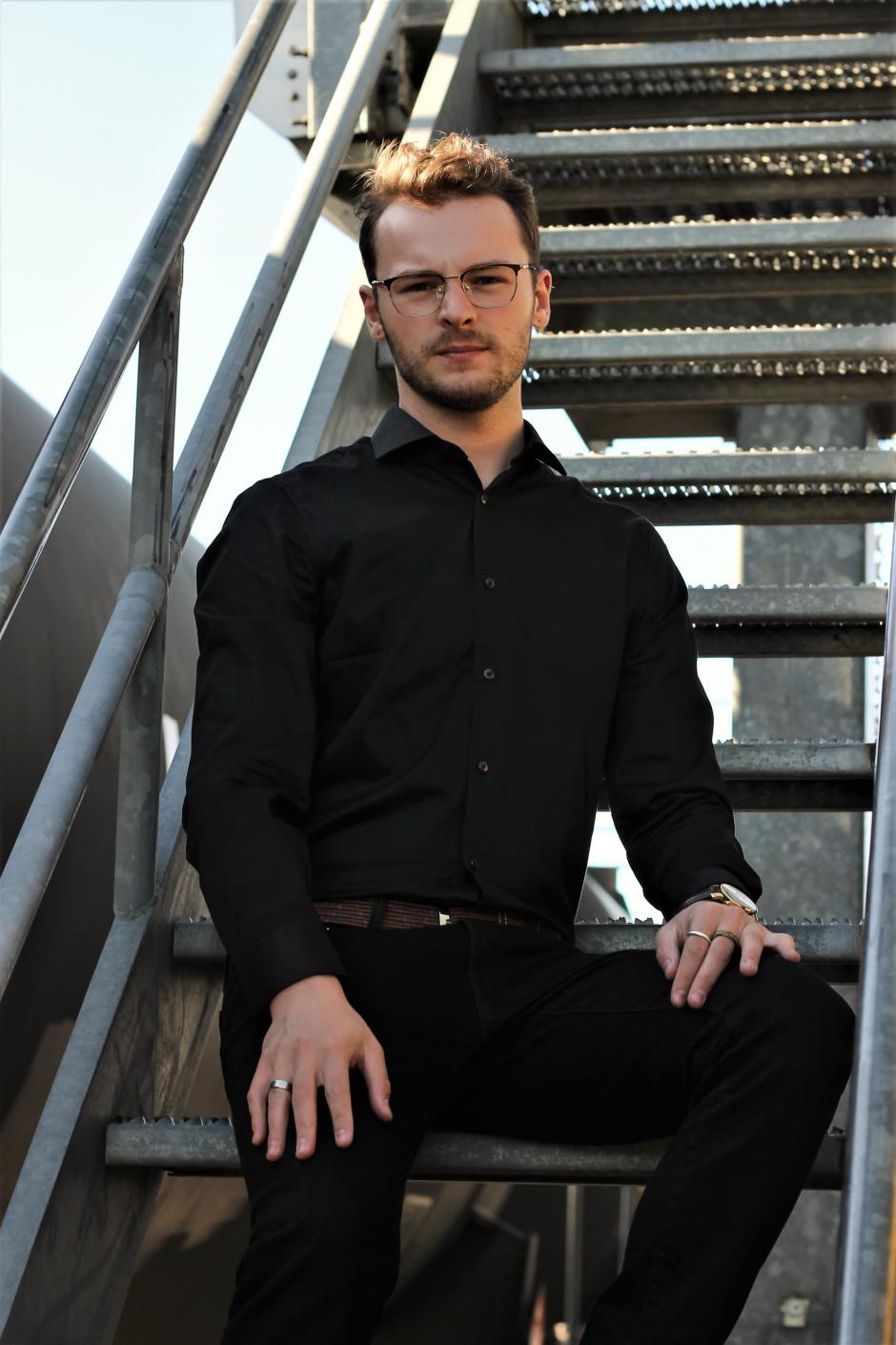 Ethan sits on metal stairs outdoors looking at the camera wearing a dress shirt and dress pants, all black.