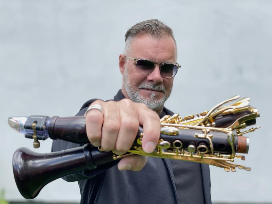 François Houle holds a deconstructed clarinet in his hand and holds it towards the camera. 