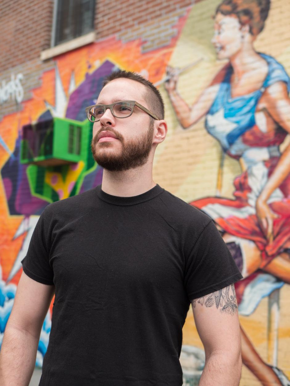 Gabriel stands in a black t-shirt looking to the left in front of a brick wall covered in paintings. 