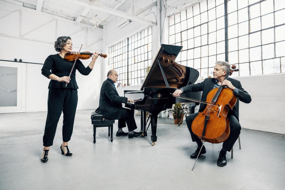 Annalee, Jamie, and Roman (left to right) are sitting in a modern white room playing their instruments (violin, piano, and cello). They are dressed in dark colours and windows are behind them.