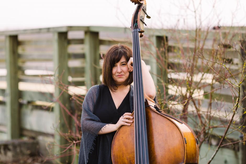 Jodi Proznick stands in front of plants and shrubs while holding her bass with one hand.