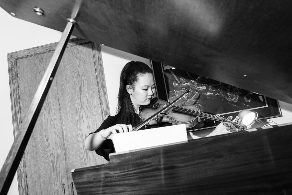 A black and white photo of Katherine playing a violin. She stands in front of a piano and the photo is shot through the piano with the lid up. There is a door behind her and a painting on the wall. 