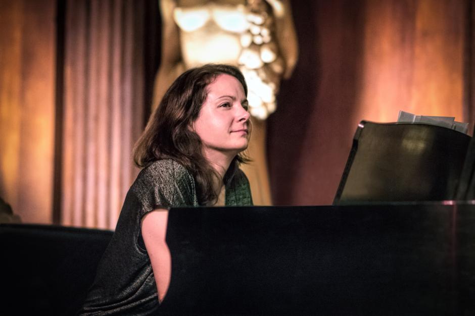 Kris Davis sits at a black piano with pink curtains and a white statue behind her.