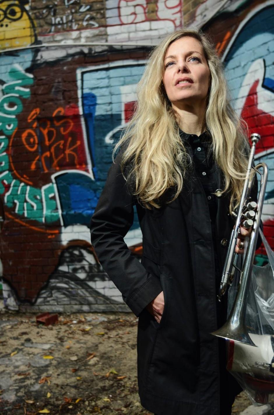 Lina stands with a trumpet in her hand. She stands against a colourful brick wall.