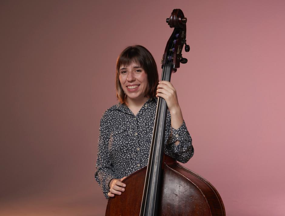 Zoe stands with a patterned shirt holding an upright bass in front of a red background. 