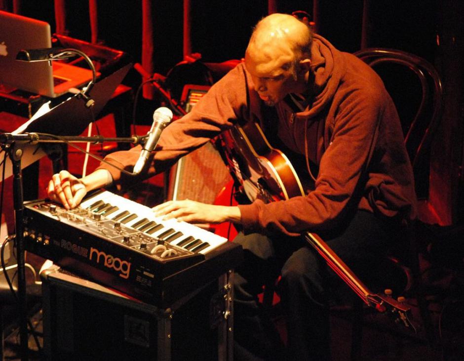 Shahzad Ismaily sits at a keyboard with a guitar in his lap. He is adjusting the keyboard and the photo is taken from above. 