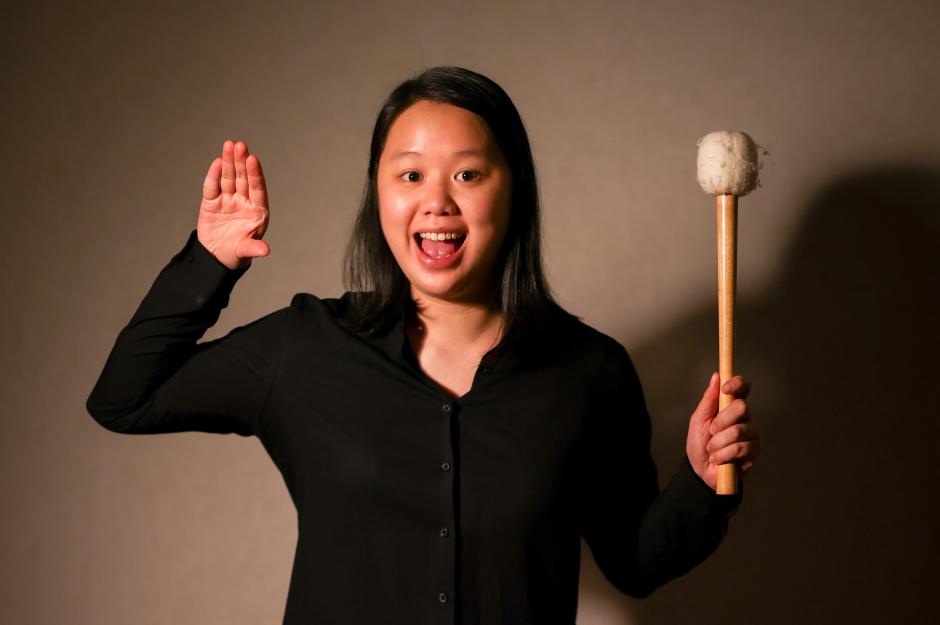 Hoi Tong Keung stands with a drum mallet and left hand out stretched. She smiles in front of a grey background. 