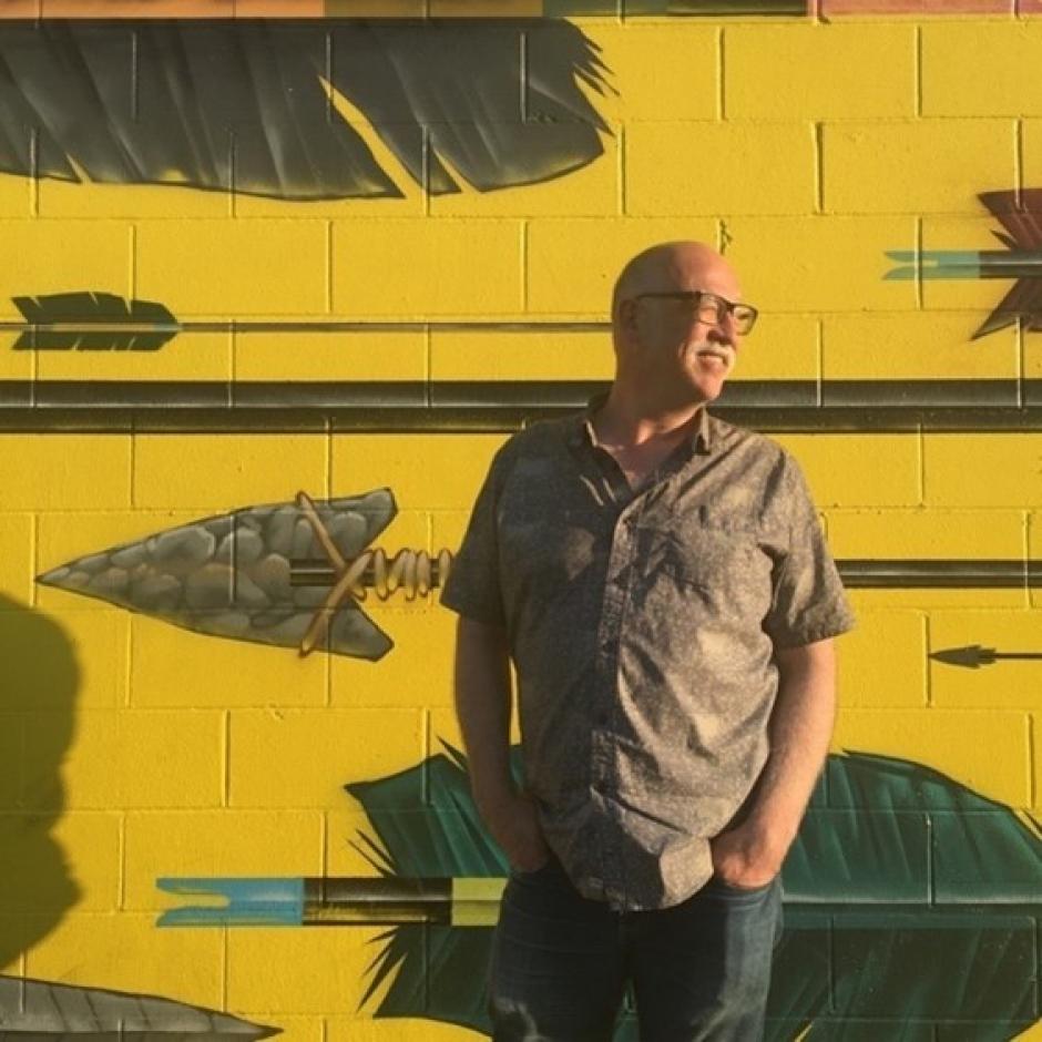 William Tennant, with his hands in his front pockets, leans against a bright yellow and green mural.