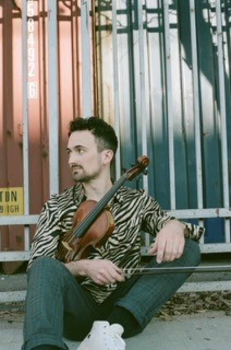 Michael sits in a patterned shirt in front of shipping containers holding a violin. 