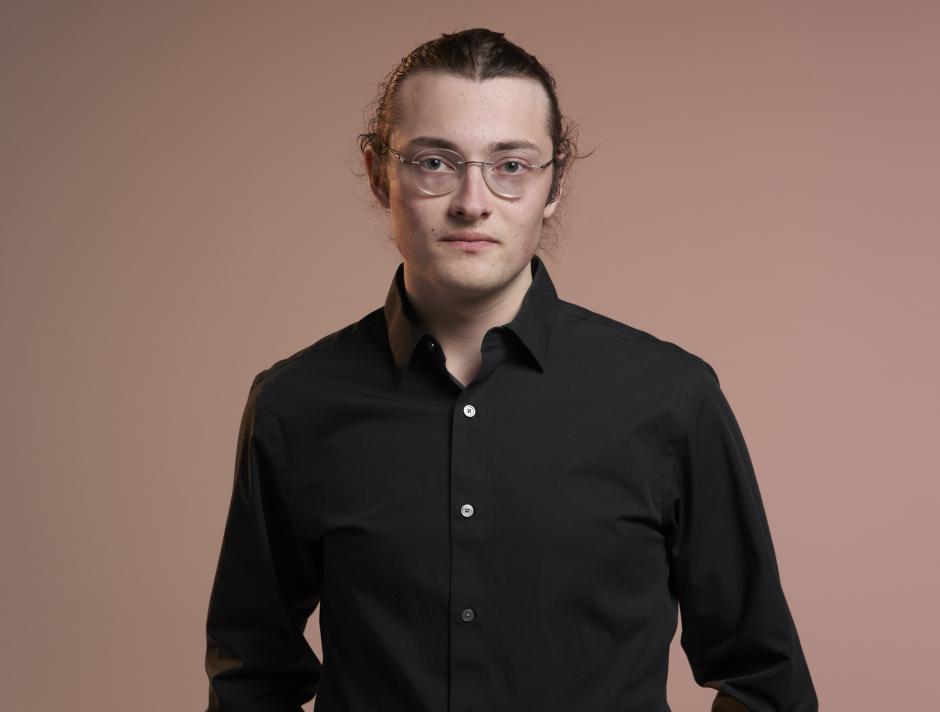 Benjamin is pictured here in front of a red background. He is wearing a black shirt and glasses. 