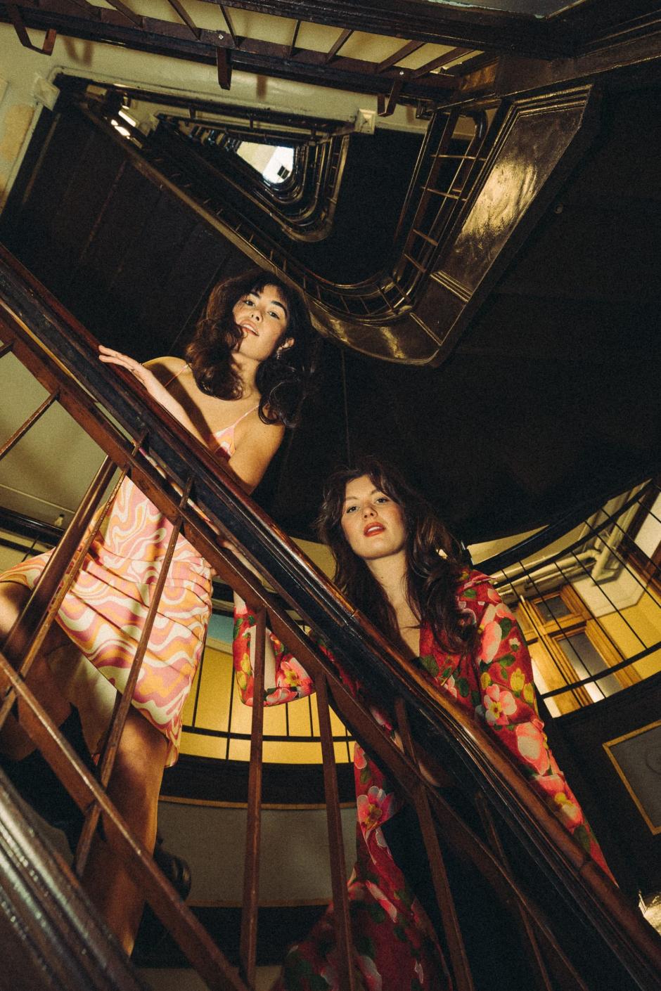 Lauren and Kate are looking down a stairwell into the lens of the camera. 