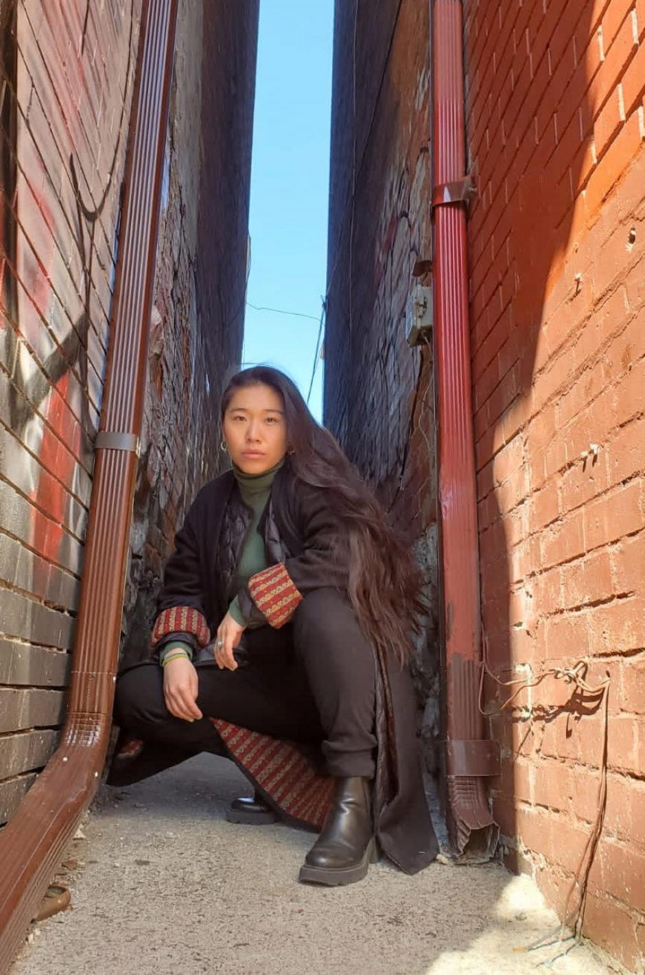Rinchen Dolma is crouching down on a narrow sidewalk between two red brick walls marked with graffiti