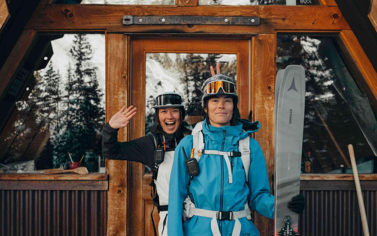 Two women with ski gears are standing in a front of a contemporary chalet door. The snowy high peaks, mountains and trees are reflecting into the windows 