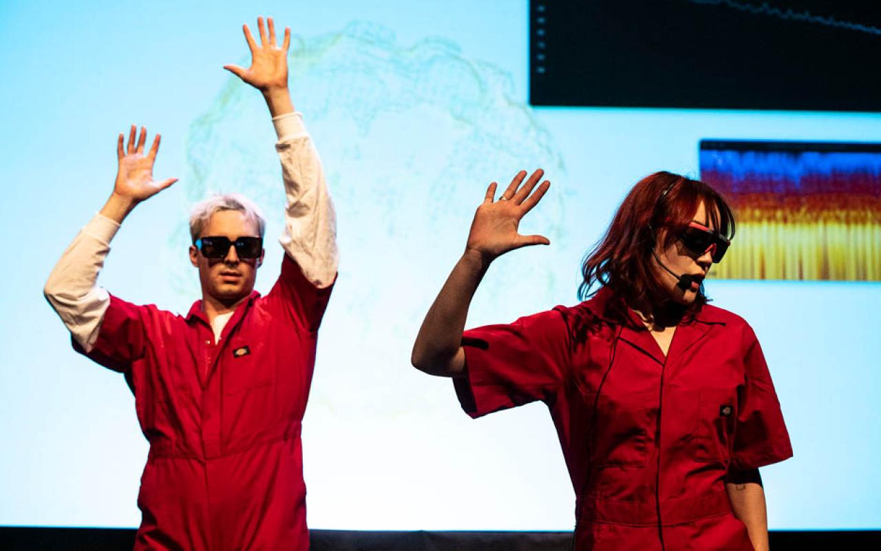 Two musicians in bright red coveralls perform in front of a blue backdrop