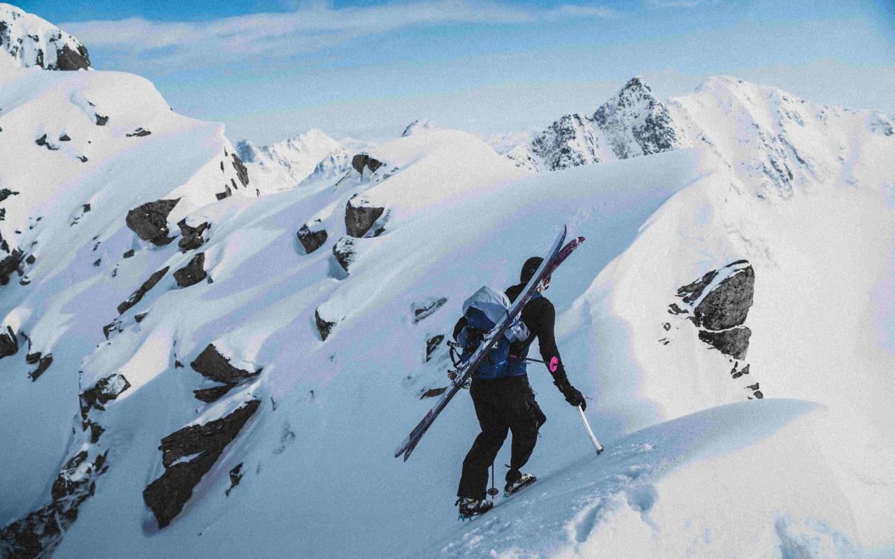 Alpine skier ascends a steep, snowy mountain ridge with tools and skis strapped to their backpack. 