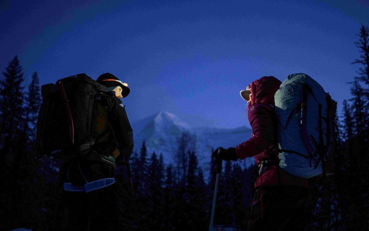 In a dark setting, a man and women with warm winter clothes, backpack, headlamp and climbing gear looking at each other, facing a snowy mountain 