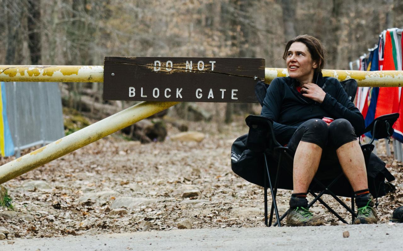 Woman wearing sport clothes and running shoes is seating in a camping chair in front of a trail head with an old wood sign indicating ''Do not block Gate''