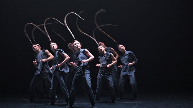 a group of 7 asian dancers on stage, wearing black clothes adn a long bird feather on their heads 