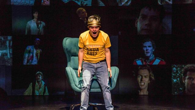 Actor wearing a gold crown and yellow T-shirt performs in front of a green armchair, surrounded by projected portraits in Teatro La Plaza’s Hamlet.