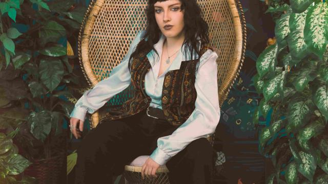 A person seated in a woven wicker chair surrounded by lush green plants and a colorful mural backdrop. They wear a white button-up shirt, patterned vest, black pants, and black shoes, with dark hair and dramatic makeup, posing confidently in a stylized indoor garden setting