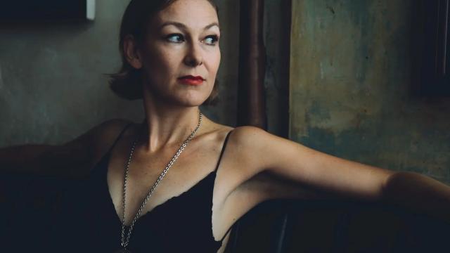 A person with short hair and red lipstick sits by a window in a black sleeveless top, gazing thoughtfully to the side in soft natural light.