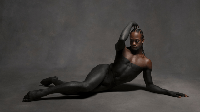 A dancer (Mykiah Goree) lies on the floor with fingertips lifted over their braids, gaze lowered. Their body is painted black to the shoulders, revealing brown skin above the paint against a gray background.