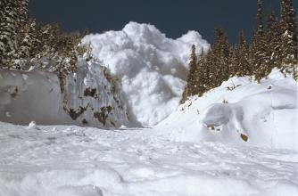 A very large avalanche is barreling down a narrow gully towards the camera, it is a blue sky above.