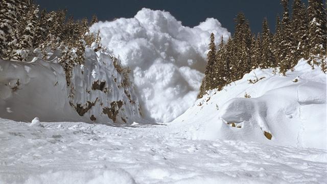 A very large avalanche is barreling down a narrow gully towards the camera, it is a blue sky above.