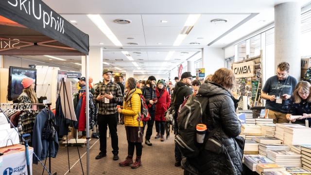 A large room is filled with numerous exhibitors interacting with the public. Books, clothing, and signage take up space on the visible tables.