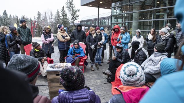 A happily animated  group dressed for winter weather stands around a gas fire pit discussing a topic.