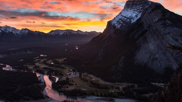 Sun rises behind Mt. Rundle as seen from Tunnel Mountain, the sky has fiery orange and bright pink clouds.