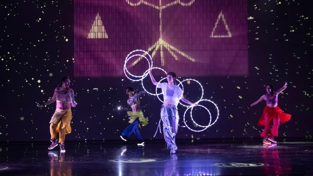 Four dancers perform on a dark stage. The centre dancer spins a chain of glowing white hoops, with star-like projections and a pink geometric backdrop behind them.