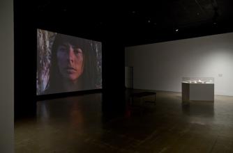 A dark gallery has a diffuse spotlight on an artistic stand, a screen in the background shows the face of an indigenous woman.